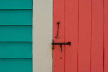 A vibrant teal green house with cream color trim and a vibrant orange shutter door. The closed door has a hook and latch attached. The locks are rusty vintage iron. The wall is narrow clapboard boards - obrazy, fototapety, plakaty