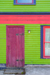 The exterior wall of a colorful door and window of an old wooden home. The purple wooden screen door has a metal latch and hinges. The window has red and blue trim against a bright lime green wall. 