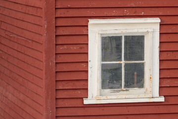 The exterior of a bright red narrow wooden horizontal clapboard wall of a house with one vinyl window. The trim on the glass panes is white. The outside boards are textured rough pine wood.  