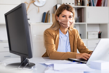 Woman in disposable face mask working in business office using laptop
