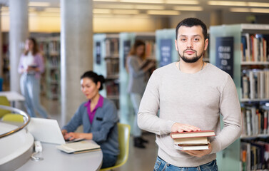 Young adult focused man holding books and looking at camera at university library, students choosing books and working on background