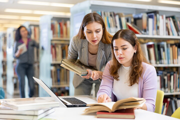 Two young positive women working together on joint project in library, finding information in books and Internet..