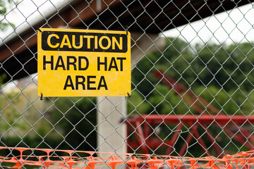 Yellow caution hard hat area sign on a chain link fence with a newly constructed bridge and crane in the background.
