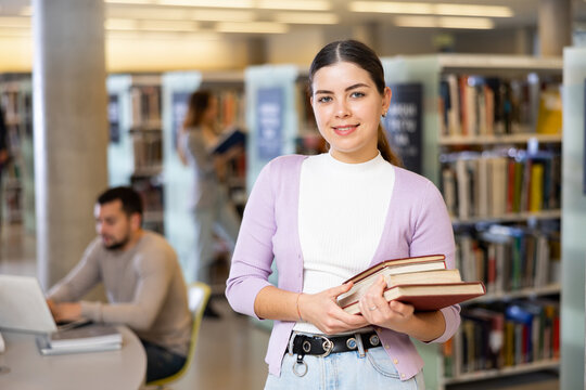 Portrait of young woman holding books, looking at camera and smiling at library