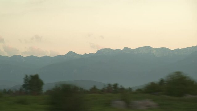 Car passes by trees and fields against high mountains in Croatia. Landscape of high mountain peaks seen from window of car driving fast after sunset