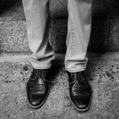 Man's legs and feet wearing cap toe shoes outside a doorstep on a New York City street. Black and...