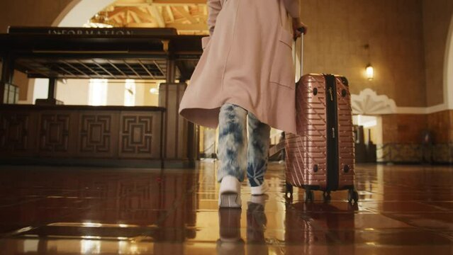 Female in long casual cardigan and white shoes with luggage on wheels walking by train station. Back view slow motion traveler woman looking for departure gates, Union Station Los Angeles USA Apr.2022