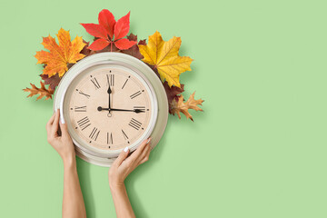 Female hands with clock and autumn leaves on color background. Daylight saving time
