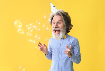 Happy senior man with big head and soap bubbles celebrating Birthday on orange background