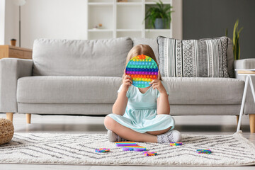 Little girl sitting on floor and holding multicolored pop it fidget toy in room - obrazy, fototapety, plakaty