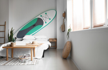 Comfortable bed and sup surfing board on light wall in room interior