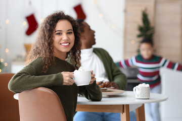 Young African-American woman with cup of hot chocolate and her family at home on Christmas eve