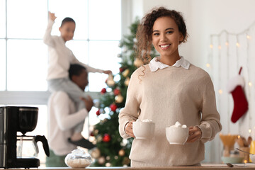 Young African-American woman with cups of hot chocolate and her family at home on Christmas eve