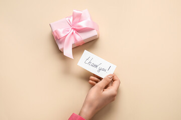 Female hand holding sheet of paper with text THANK YOU and gift box on beige background