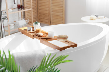 Wooden bathtub tray with different supplies and cosmetic products in light bathroom