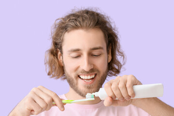 Young man applying tooth paste onto brush on lilac background, closeup