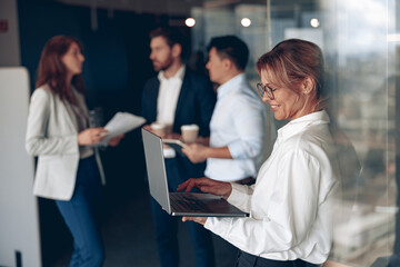 mature confident businesswoman at office with group of colleagues on background, working on laptop.