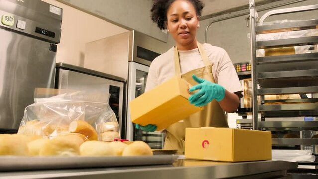 Bakery startup small business delivery. One African American female cook is packing handmade and fresh-baked bread and pastries in boxes and sending for online customer purchases in culinary kitchen.
