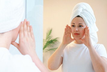 Young woman with a white towel on her head doing self-massage of the face near mirror.