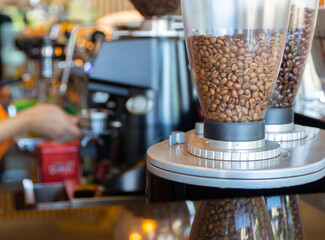 Close up shot with copy space of coffee beans in dispenser machine placing on table top in coffee shop with blur background of coffee preparation by barista for background of beverage business.