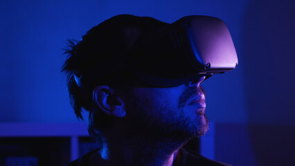 Close-up of a bearded man wearing VR glasses in a dark room with blue light. There is a room flower in the front captivity. VR helmet for entertainment, learning and gaming.
