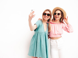 Two young beautiful smiling brunette hipster female in trendy summer dresses. Sexy carefree women posing near white wall. Positive models having fun. Cheerful and happy. In hats. Taking selfie photos