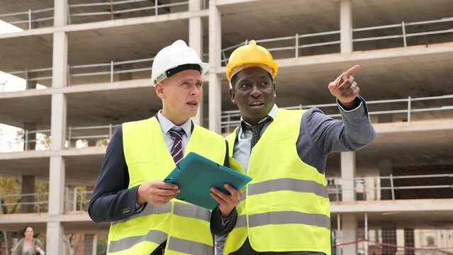 Two architects, Caucasian and African-american men in warnvests, standing on building site and discussing project documentation. One man pointing with his finger.