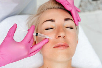 Professional cosmetology. Close-up portrait of beautiful adult woman getting injection in the cosmetology salon. Doctor in medical gloves with syringe injects filler in nasolabial fold