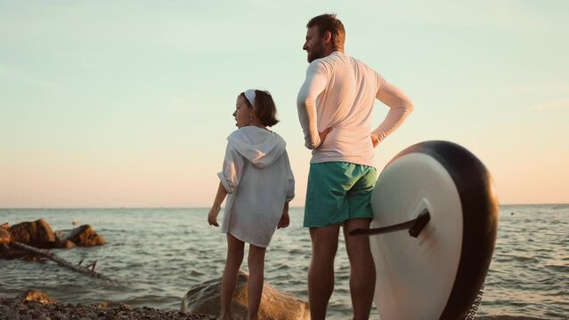 Father and daughter stands on the ocean's beach with a sup board and talking to each other. Back view. Slow motion.