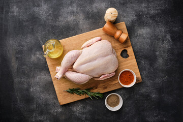 A whole raw chicken resting on a wooden board against a dark background. Preparing raw chicken. Top view © FoodAndPhoto