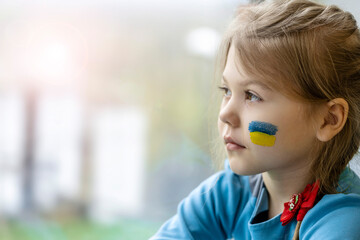 Portrait of an 8-year-old girl with a Ukrainian flag on her face, sitting by the window.