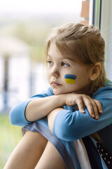 Portrait of an 8-year-old girl with a Ukrainian flag on her face, sitting by the window.
