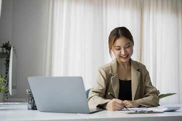 Beautiful female employee using laptop computer and making important notes on notebook.