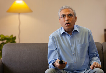 Shocked senior man sitting on sofa in the living room and watching television.