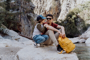 School boy and his dad with yellow backpack sits on a riverside rock in the canyon with mountain cliffs in the background. Kid child and father taking a rest on a boulder near mountain river. - obrazy, fototapety, plakaty