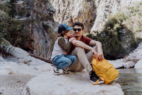 School boy and his dad with yellow backpack sits on a riverside rock in the canyon with mountain cliffs in the background. Kid child and father taking a rest on a boulder near mountain river. - Powered by Adobe