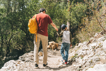 Rear view of tourists school boy and his dad walking a stone footpath in spring forest. Child kid and father wearing casual clothes and yellow backpack while hiking in summer greenwood leaf forest.