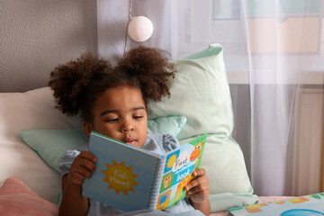 African American girl reading book on bed at home
