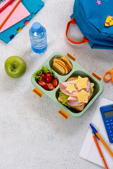 Healthy school lunch box with sandwich and salad on the workplace near backpack. School supplies, books, apple and a bottle of water.