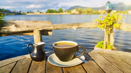 A casual morning cup of coffee with creamer served on a rustic wooden table beside a lake on a sunny day, a peaceful and relaxing setting on a summer vacation. Shot in Cambodia.