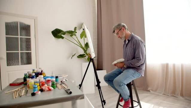 Man is sitting chair and drawing on an easel with brush holding palette in hands, there are many cans of paint and brushes on table, side view. Concept of recreation