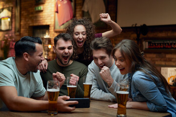Group of cheerful soccer fans having beer and watching a match on smartphone in a pub