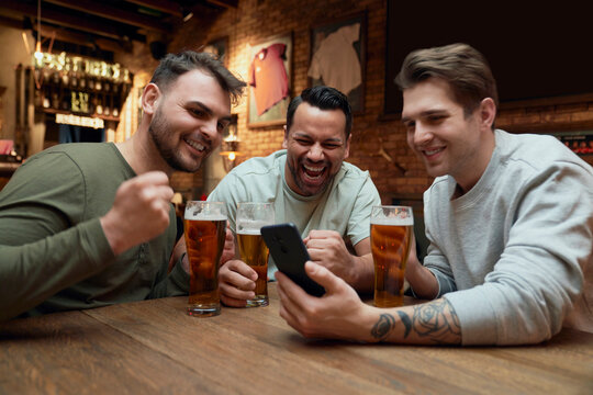 Three cheerful male soccer fans having beer and watching a match on smartphone in a pub