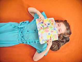 Girl covering mouth with book lying on orange background