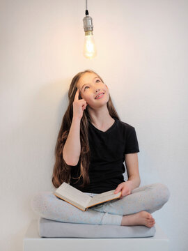 Thoughtful girl with book sitting below light bulb in front of white wall
