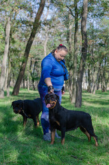 A woman in a blue sports uniform and two black dogs standing on green grass. Handler and adult female Rottweiler.