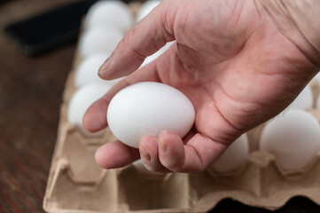 A man is holding a chicken egg in front of an egg tray. Adult male holding a white raw egg. Open recycled tray. Selective Focus.