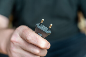 A mature man's hand is holding an electrical plug. A black plastic AC power plug.