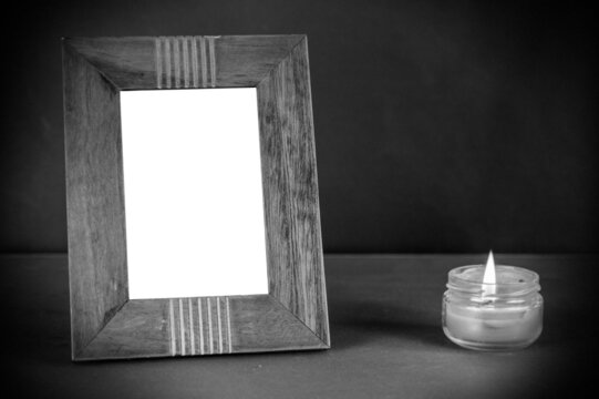 A wide brown empty photo frame set on a black surface next to a burning candle. A lit candle in a clear glass jar. Vertical placement of rectangular photo frame.