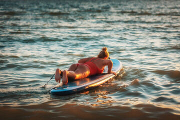 Adult plump woman in a red swimsuit, swimming in the sea on a sup board. Copy space. The concept of active recreation at sea and sports